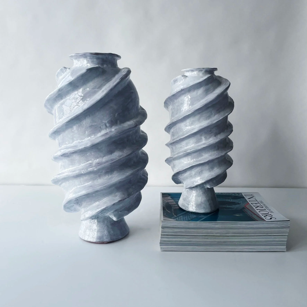 The Ithaka Vase by Ellen Robinson Studio, featuring a white glaze with spiral, textured designs, is displayed on a stack of magazines alongside another sculptural vase, both set on a white surface.