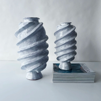 The Ithaka Vase by Ellen Robinson Studio, featuring a white glaze with spiral, textured designs, is displayed on a stack of magazines alongside another sculptural vase, both set on a white surface.