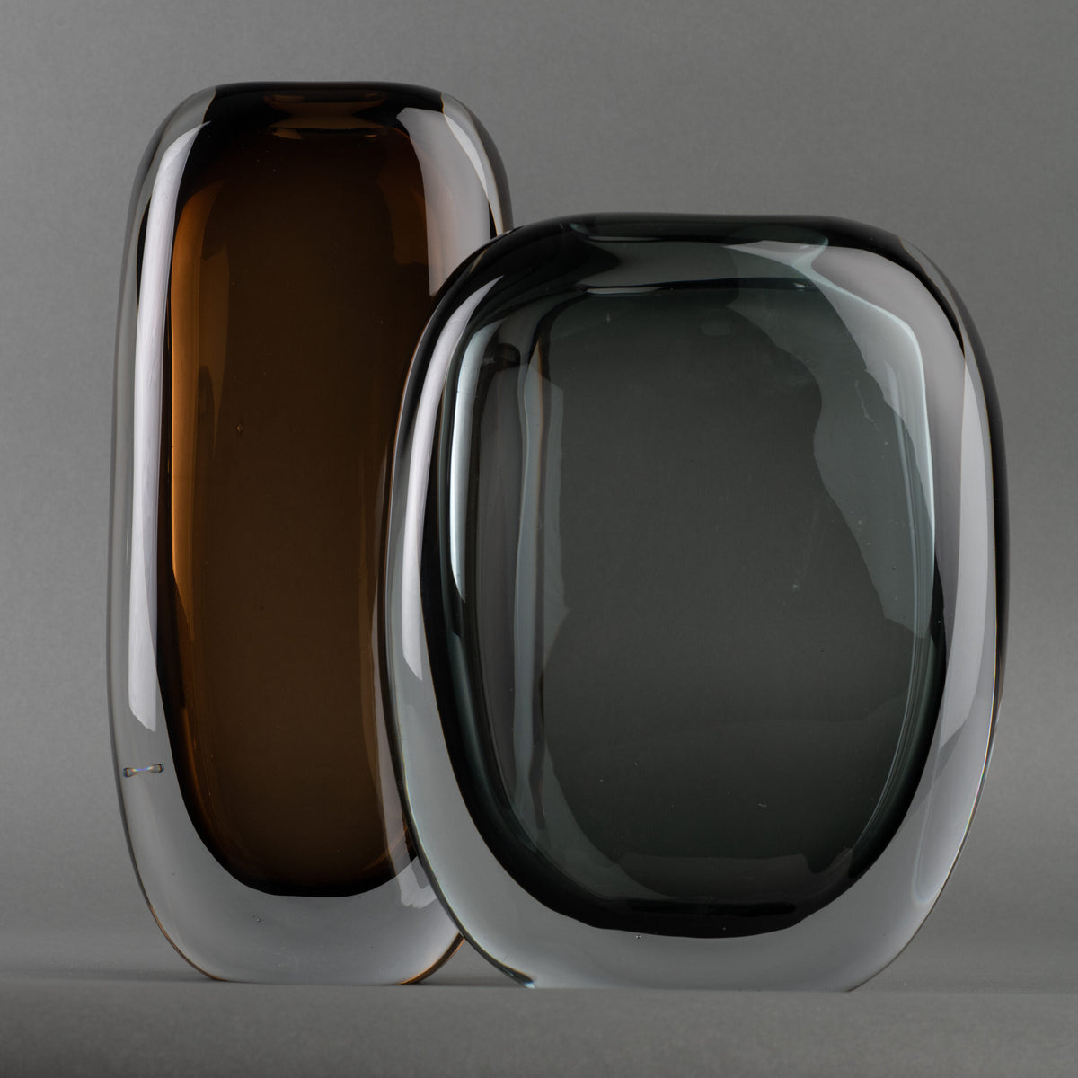 Two modern oval glass sculptures stand side by side against a gray background—one an amber brown Paul Vase by FlowDecor Inc., the other a smokey grey vase—both with clear edges, ideal for elevating any home’s interior décor.