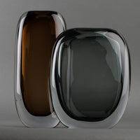 Two modern oval glass sculptures stand side by side against a gray background—one an amber brown Paul Vase by FlowDecor Inc., the other a smokey grey vase—both with clear edges, ideal for elevating any home’s interior décor.