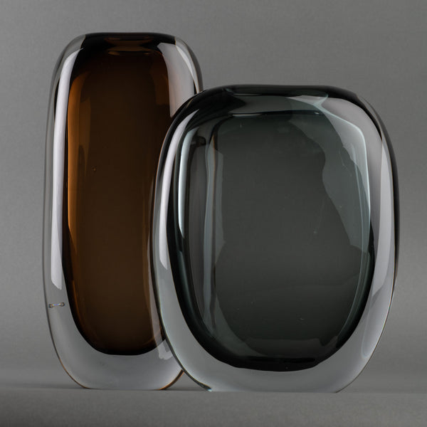 Two modern oval glass sculptures stand side by side against a gray background—one an amber brown Paul Vase by FlowDecor Inc., the other a smokey grey vase—both with clear edges, ideal for elevating any home’s interior décor.