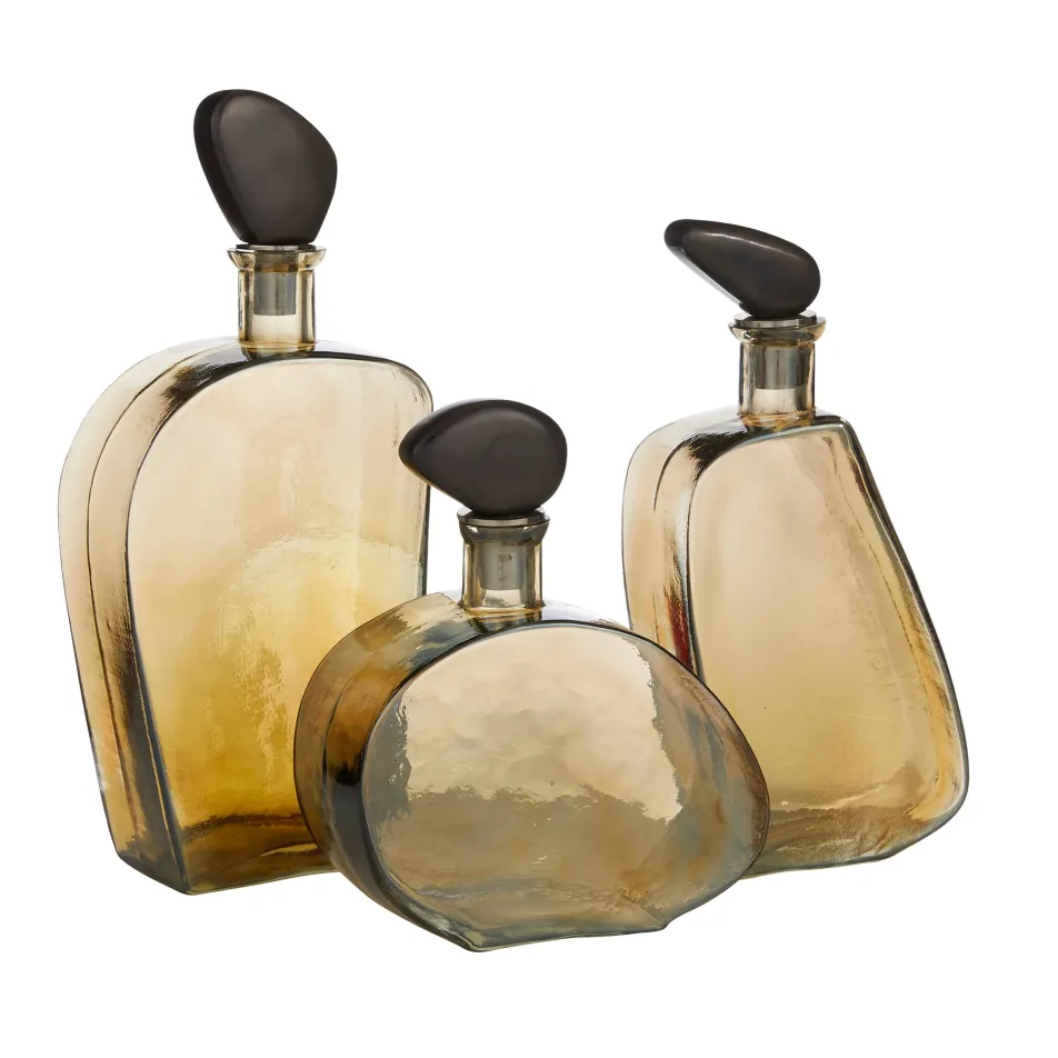 Three amber-colored Calhoun Decanters by Arteriors feature unique, irregular hammered glass forms with dark rounded stoppers, grouped together on a white background.