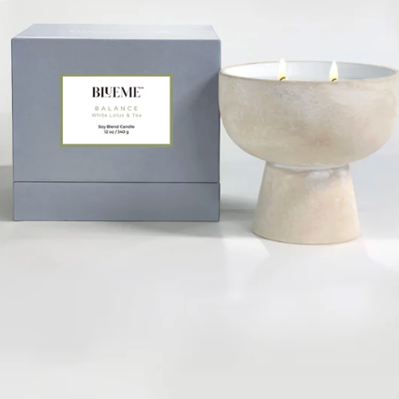 A pale ceramic Balance Candle by Blueme, made with a soy wax blend and dual wicks, sits beside a light gray box—an inviting wellness fragrance featuring White Lotus & Tea for your space.