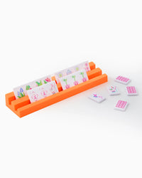 A vibrant orange tile holder from the Oh My Mahjong Mahjong Debutante Travel Tile Set displays illustrated tiles, some featuring flowers and animals, with extra tiles scattered nearby on a white background.