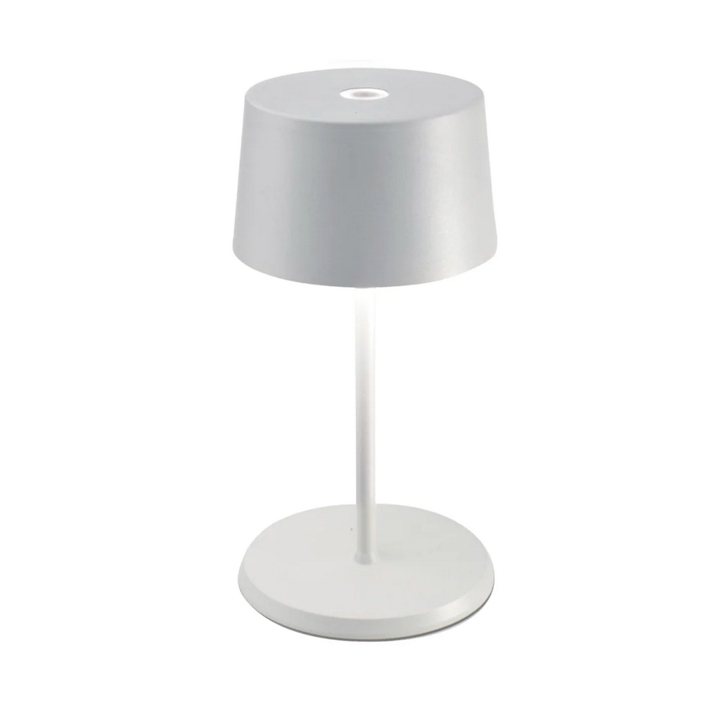 The Olivia Mini Table Lamp by Zafferano is a modern, minimalist white lamp featuring a round base, slender stem, and cylindrical shade. Its dimmable LED offers convenient touch control for easy lighting adjustments.