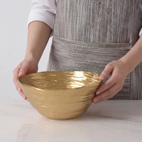 A person in a gray striped apron holds the Dorrato Gold Bowl - by PAMPA BAY, a large textured matte gold bowl, on a white surface.