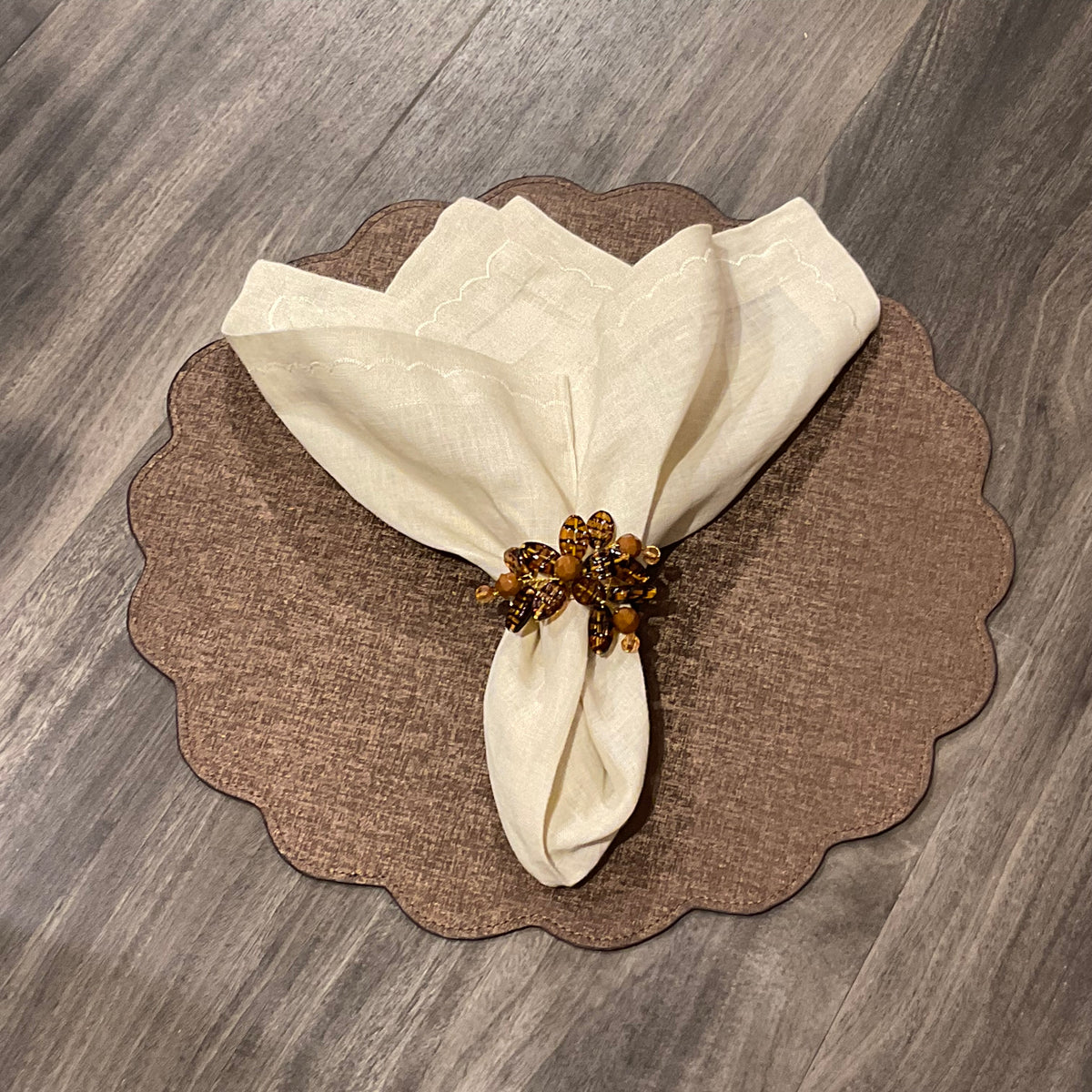 A cream-colored cloth napkin with a beaded ring rests on the Deborah Rhodes Victoria Round Scallop Placemat Set of 4, bringing elegance and easy care to your wooden table.