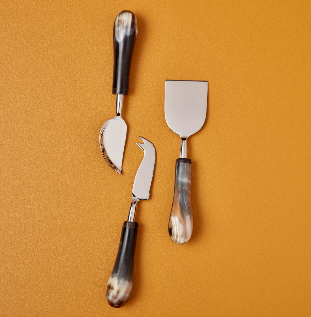Three cheese knives from the Be Home Tusk Handle Cheese Set, with gleaming metal blades and rich brown marbled handles, are arranged vertically against a solid mustard-yellow background—a sophisticated showcase of modern luxury ideal for elevating your next cheese board.