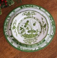 Two Faenza Melamine Dinnerware plates from Beatriz Ball, featuring green and white botanical and geometric designs, are stacked on a wooden table. Greenery with red berries appears partially in the upper left corner. Set of 4.