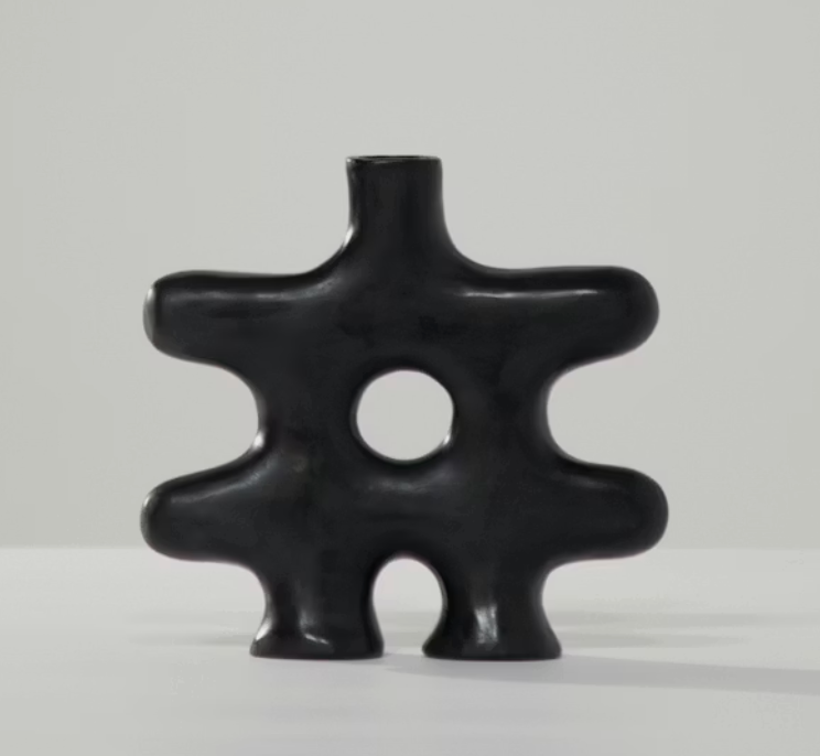The Nedjma Vase - Black by Schwung Design is a modern black abstract ceramic vessel featuring a central hole and four rounded protrusions, evoking sculptural curves or an organic gear-like form. Crafted with artistry inspired by handmade Mediterranean techniques, it sits against a light gray background.