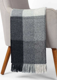 The Sloane Alpaca Throw by Alicia Peru Home, featuring a large black, gray, and white plaid pattern with tassel trim, is draped over the arm of a light gray armchair with wooden legs.