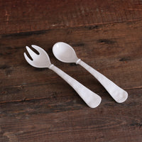 The Beatriz Ball Vida Havana Salad Servers - White, featuring one forked and one spoon-ended melamine server, are displayed side by side on a rustic wooden surface. These servers are decorative, practical, and dishwasher safe.