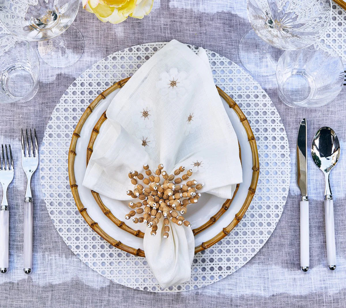 A table setting featuring Kim Seybert's Reed Placemats Set of 4 in White, a decorative plate, bamboo-rimmed plate, beaded ring napkin, glassware, and silver cutlery—yellow flowers complete this chic look for outdoor entertaining.