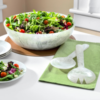 The Eddy Resin Bowl With Servers by Inspired Generations—featuring a large marble-patterned design—holds a fresh green salad with tomatoes atop a mat. Nearby, matching marble servers, a small coordinating plate, and a green napkin rest on the white table, making this stylish set an ideal housewarming gift.