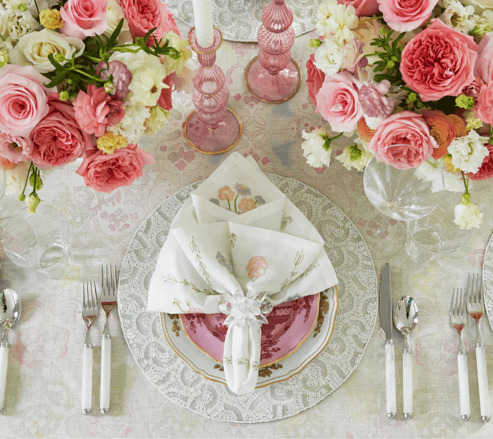 A beautifully set table features the Kim Seybert Giverny Tablecloth in Mint & Multi, pink roses, a floral centerpiece, pink glass candle holders, and place settings with embroidered napkins and delicate china.