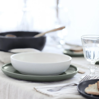 A white bowl from the Casafina/Costa Nova Pacifica Dinnerware Artichoke collection rests on a green plate atop a set dining table featuring luxury tableware, complemented by a glass of water, cutlery, and a piece of bread. A black pan and a blurred background are also visible.