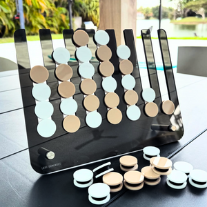 The Connect Four El Cuatro game by Luxe Dominoes, featuring chic beige and white pieces, is elegantly arranged on a black table outdoors. Extra pieces are on hand, while the backdrop of trees, water, and patio furniture creates an inviting atmosphere—perfect for challenging friends to see who can get four in a row first.
