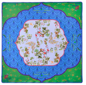 The Oh My Mahjong "Mahjong Mats" silk scarf features a central white area with small Asian-style houses, trees, and flowers, bordered by blue scalloped patterns and a green edge with decorative details and text.