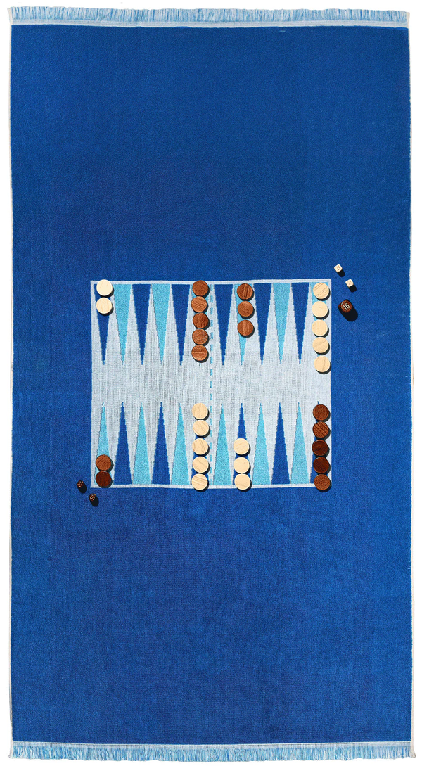 The Southampton Beach Backgammon by G. McGee is a plush blue cotton terry towel with white fringe, featuring a woven backgammon board of blue and white triangles and includes brown and beige wooden playing pieces.