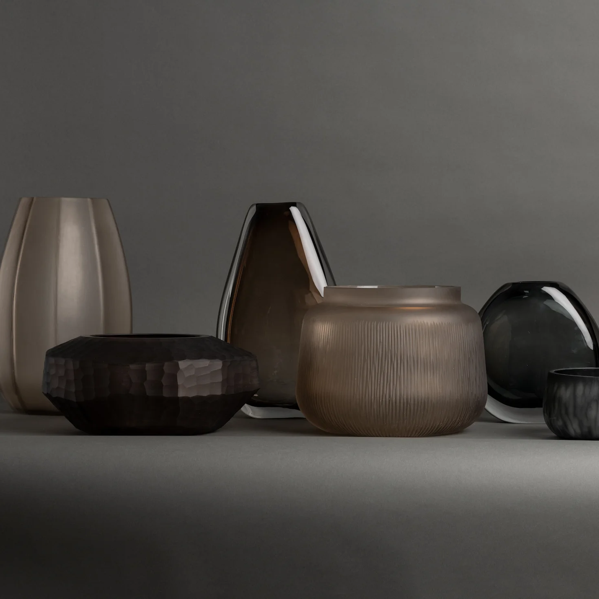 A set of six decorative vases, featuring the Paul Vase by FlowDecor Inc. and a smokey grey glass vase in assorted shapes, sizes, and muted tones, displayed on a gray background—perfect for enhancing your home décor.