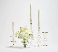 A group of clear glass candlesticks with white candles and a matching Kim Seybert Scallop Bud Vase in clear, elegantly holding white flowers—all featuring gold trim—are displayed on a white background.