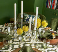 Elegant table set with green napkins, a patterned tablecloth, clear mouth-blown glassware, white candles in glass holders, and yellow flowers arranged in hand-crafted vases—including the Kim Seybert Scallop Bud Vase - Clear. Books are visible on a green shelf in the background.