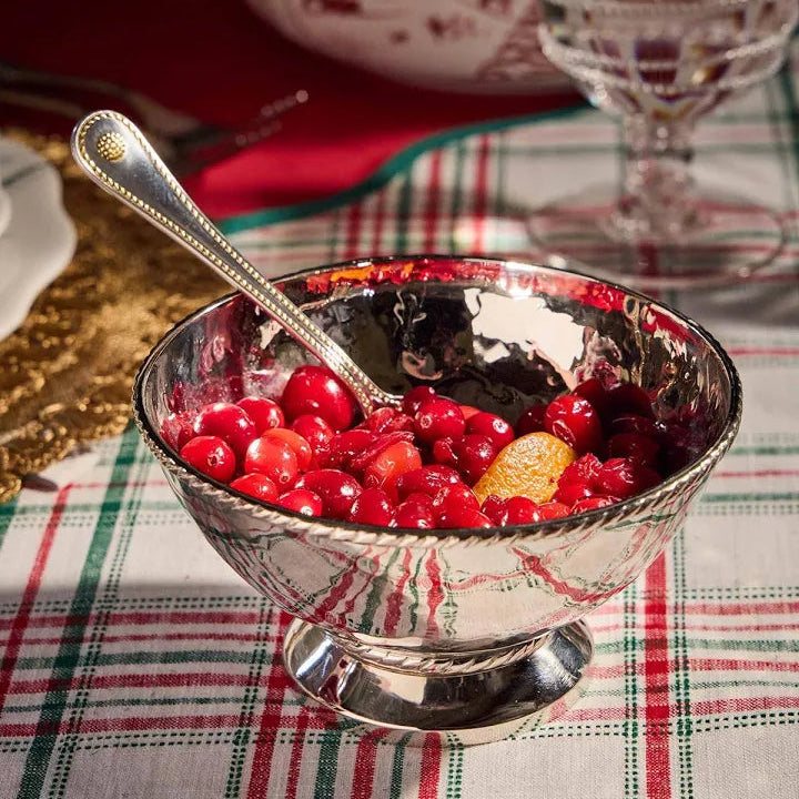 The Juliska Graham Cocktail Bowl, crafted from hammered stainless steel, is filled with cranberry sauce and orange slices on a plaid tablecloth, with a silver spoon resting inside.