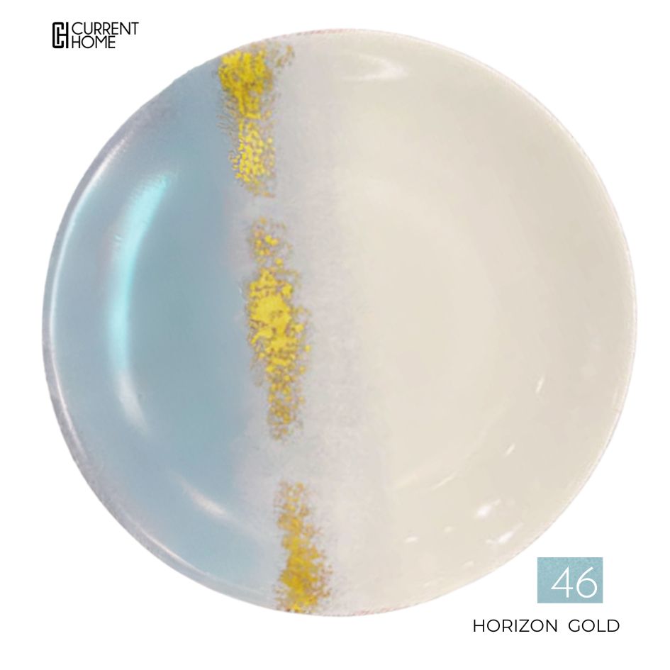 A round, hand-painted Marie Daâge Horizon Gold Dinnerware plate features a blue left side, white right side, and a gold-white vertical stripe. "Current Home" is in the top left; "46 HORIZON GOLD" in the bottom right. Made in France.
