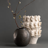 The Yvette Vase by FlowDecor Inc.: a dark, round ceramic vase with textured lines and flowering branches, paired with a tall off-white vase featuring sculptural, petal-like accents, displayed on a gray background.