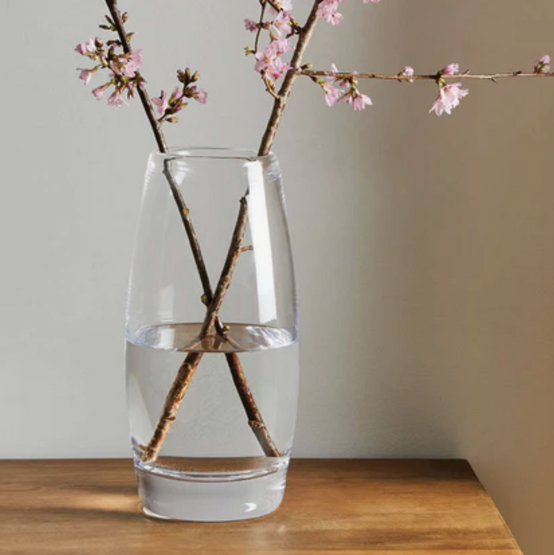 A clear, lead-free Simon Pearce Addison Vase holds water and two intersecting pink cherry blossom branches, set on a wooden surface against a neutral wall.