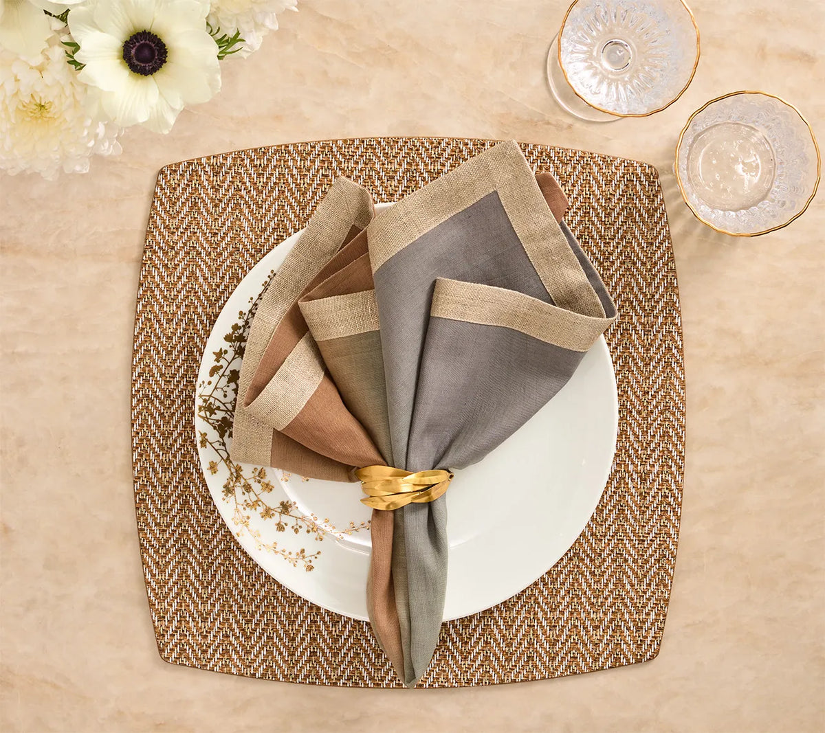 A square Kim Seybert Twill Placemat Set of 4 - Brown & White topped with a white plate, a gold-toned napkin ring, and folded napkins in brown, beige, and grey.