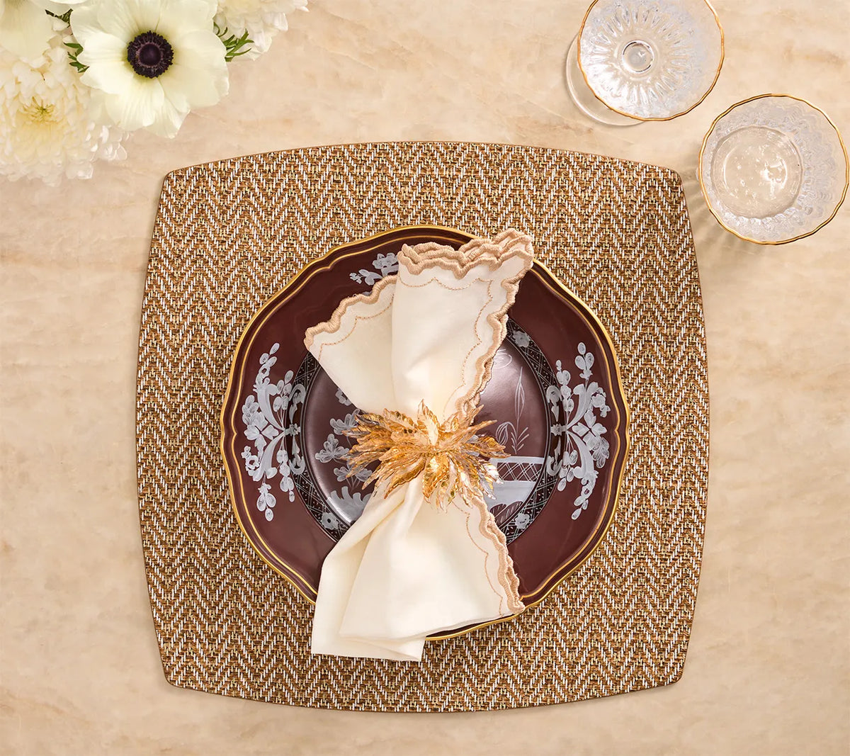 A square, brown and white Twill Placemat Set of 4 - Brown & White topped with a dark plate, a cream-colored napkin, and a metallic floral napkin ring.
