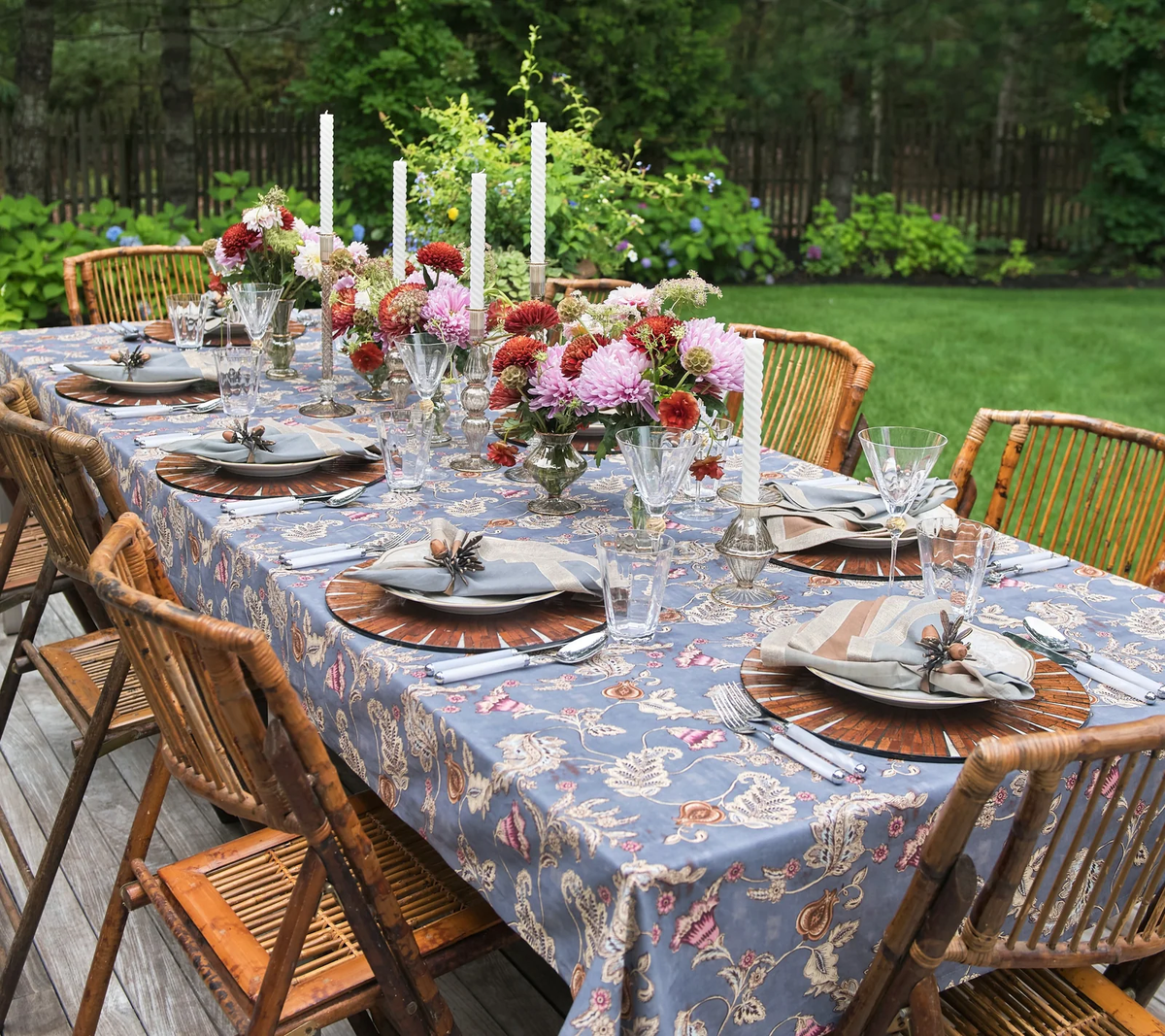 An outdoor dining table set with floral arrangements, candles, glassware, plates, and bamboo chairs is elegantly styled with the Kim Seybert Paisley Vine Tablecloth in Blue & Gray (54x126″) draped across the surface. The sophisticated arrangement sits on a deck overlooking a green lawn and trees in the background.