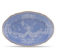 The Oriente Italiano Pervinca by Ginori 1735 is a blue oval ceramic platter with scalloped edges, a white floral and landscape pattern, and a 24-karat gold rim, evoking classic elegance.