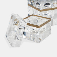 The Gemma Box by Luho is a clear, square acrylic decor box with a removable lid, rounded indents, and a gold metal accent around the opening. This modern luxury piece is partially reflected on a white surface.