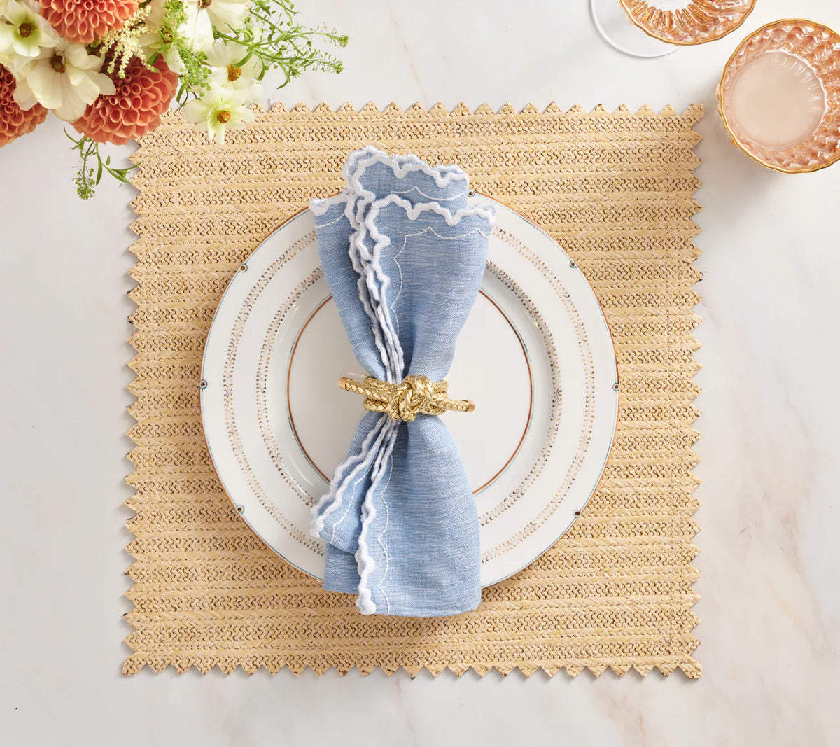 A Vista Reversible Placemat set in Natural & Brown by Kim Seybert holds a white plate with gold trim, a blue napkin with ruffled white edge in a gold ring; flowers and pink glassware are nearby.