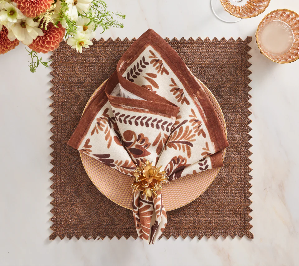 A place setting featuring a Kim Seybert Vista Reversible Placemat in Natural & Brown, a rose gold plate, a folded white napkin with a brown floral pattern, and a gold flower napkin ring.