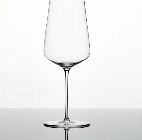 A clear, empty Zalto Universal Glass by Grand Cru Selections / Zalto, made from lead-free crystal with a long stem and wide bowl, rests on a reflective surface against a plain white background.