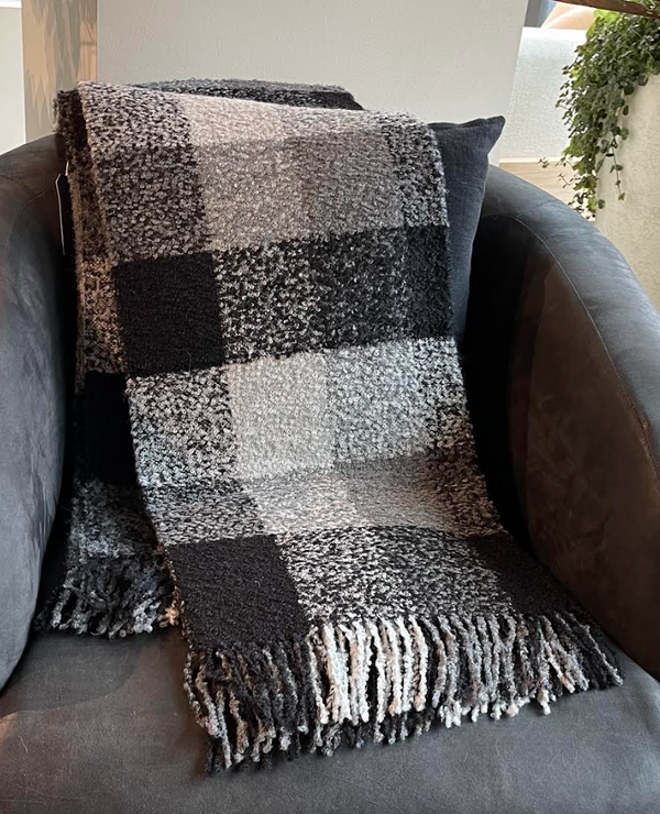 The Alicia Peru Home Bristol Boucle Throw in Grey / Black, featuring a checkered pattern and fringed ends, is draped over a dark gray armchair. A cushion rests behind the throw, while a hanging plant can be seen in the background.