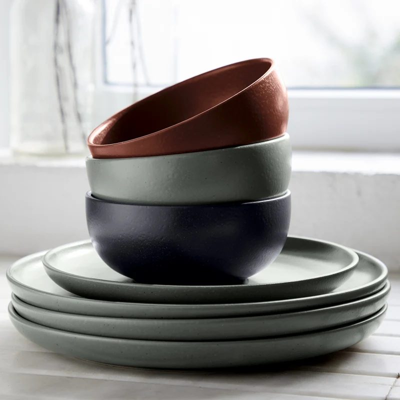 Four stacked ceramic bowls in muted green, black, and terracotta from the Casafina/Costa Nova Pacifica Dinnerware Artichoke collection rest atop a stack of four matching green plates on a white surface near a window, bringing understated luxury to the table setting.
