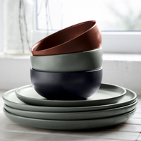 Four stacked ceramic bowls in muted green, black, and terracotta from the Casafina/Costa Nova Pacifica Dinnerware Artichoke collection rest atop a stack of four matching green plates on a white surface near a window, bringing understated luxury to the table setting.