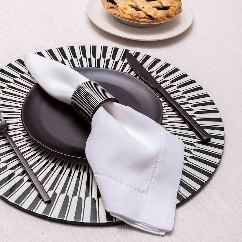 A place setting features a white napkin on a black plate, set atop the Manhattan Lacquer Placemat by Von Gern Home in a modern geometric style, with a piano key charger and a small pie in the background.