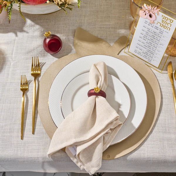 The table is elegantly set with gold cutlery, white plates arranged on Waterdale Collection’s Leather Apple Charger Set of 4 in silver, a beige napkin adorned with a pomegranate napkin ring, a red glass, and a holiday menu card featuring Hebrew text atop a light tablecloth.