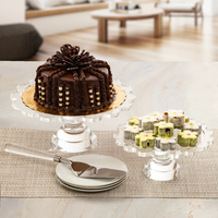 A chocolate cake with decorative icing and striped accents sits on the Coco Scalloped Cake Server by Inspired Generations, next to assorted pastries arranged on a clear pedestal server, with plates and utensils set out on a light table in a modern room.