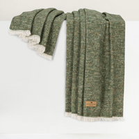 A Cinder Mist Throw by Lands Downunder, crafted in a modern style with green woven fabric and white fringe, is draped over a white surface. A brown rectangular label is attached to one corner of the blanket.