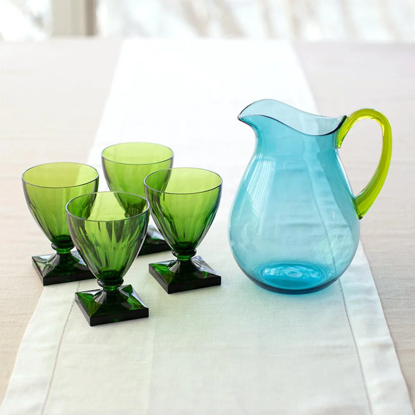 A Caspari Acrylic Handled Pitcher in turquoise & green sits beside four green goblets on a white table runner atop a light table with a shatter-resistant surface and acrylic dinnerware.