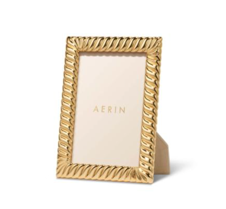 The Aerin Loretta Frame Gold features a textured, twist design and stands upright on a white surface. It holds a placeholder card with "AERIN" in gold letters and fits a 4x6 photo.