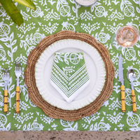 A table setting with a green and white patterned tablecloth, rattan charger, white plate, Pomegranate Napkin Set of 4 - Green by Pomegranate, bamboo-handled cutlery, and a glass of rosé wine.
