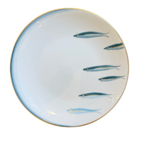The Marie Daâge Mer d'iroise 1 Dinnerware is a round white ceramic plate with a gold rim and six blue fish in two rows on the right, plus a subtle blue gradient along the left—an elegant design inspired by Limoges porcelain.