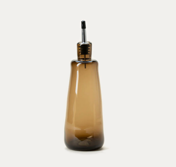 The Handblown Oil Cruet by Gary Bodker features a tall, translucent brown glass design with a sleek black pour spout, crafted for easy dispensing of oil or vinegar. This high-quality kitchen accessory, photographed against a plain white background, exemplifies exceptional craftsmanship and functional elegance.